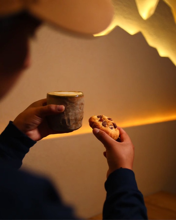 Una mano sostiene una taza de cerámica y una galleta frente a la pared de montañas iluminada